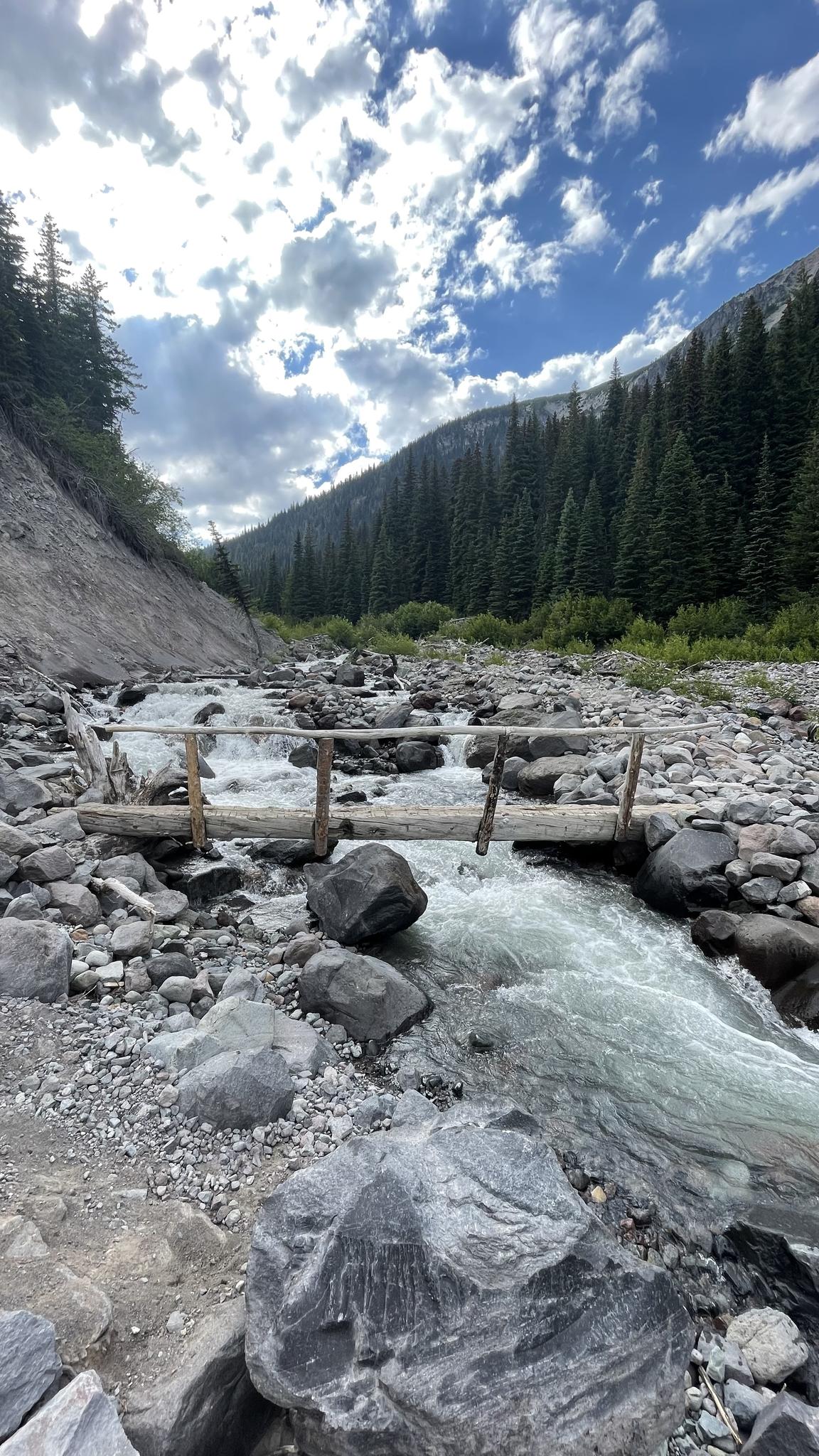 Rushing river flows over rocks, with a small wooden bridge and forested mountains.