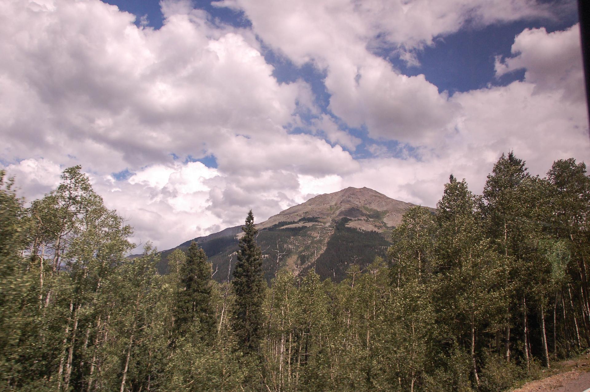 A mountain peak rises above a dense forest under a cloudy sky.
