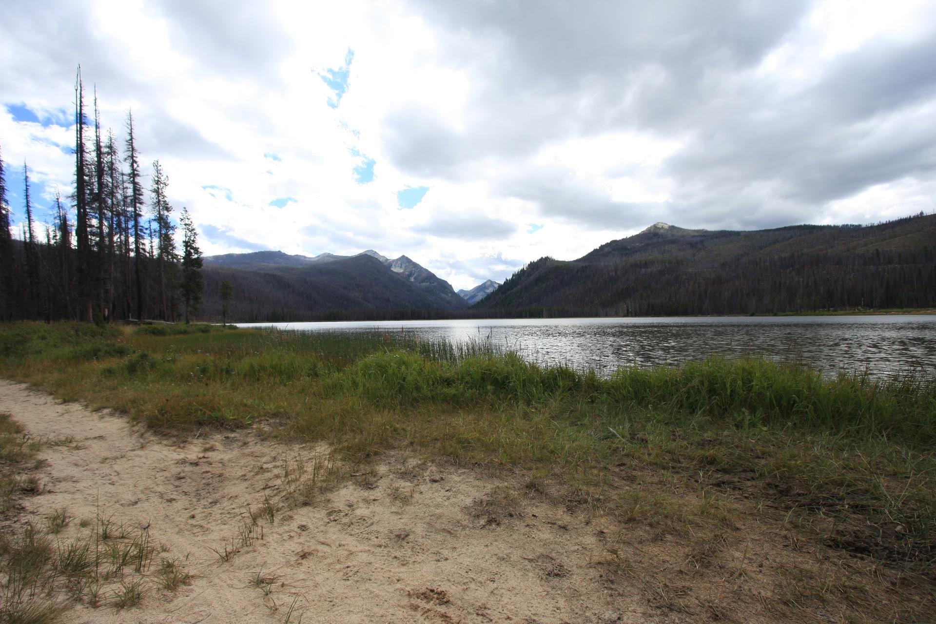 Wide lake with distant mountains under an overcast sky, sandy path in foreground.