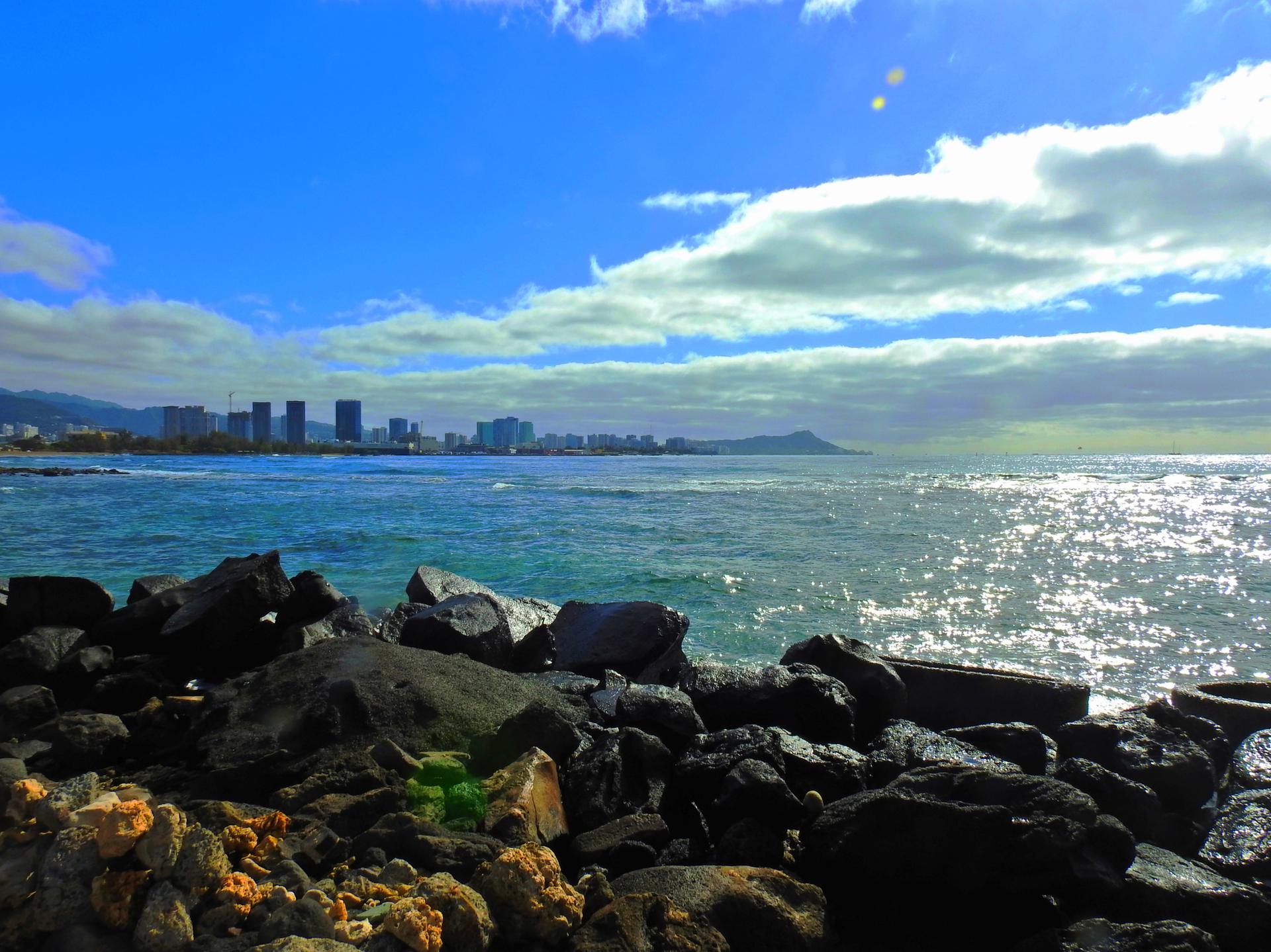 Rocky shoreline with sparkling ocean and distant city under bright sun.