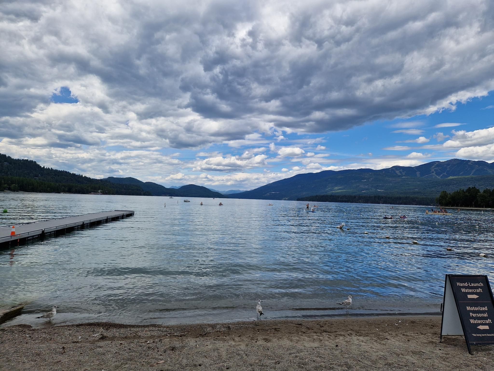 Wide lake with distant mountains, dock, sandy beach, and swimmers. - Downtown to Whitefish City Beach via Fish Trails Bike and Pedestrian Path loop