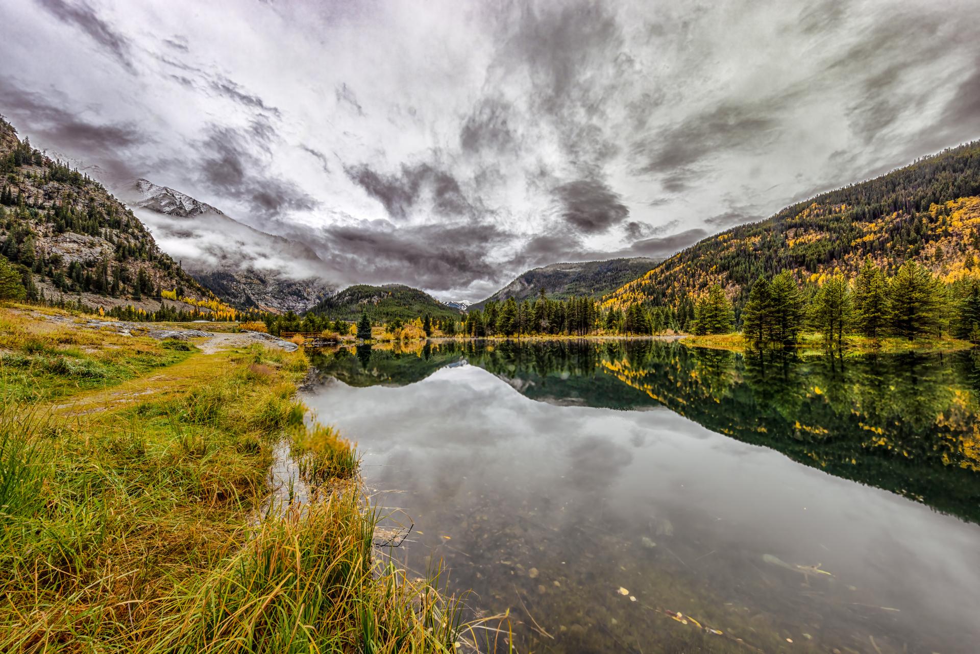 Mountain lake reflecting cloudy sky and autumn trees with grassy shore.