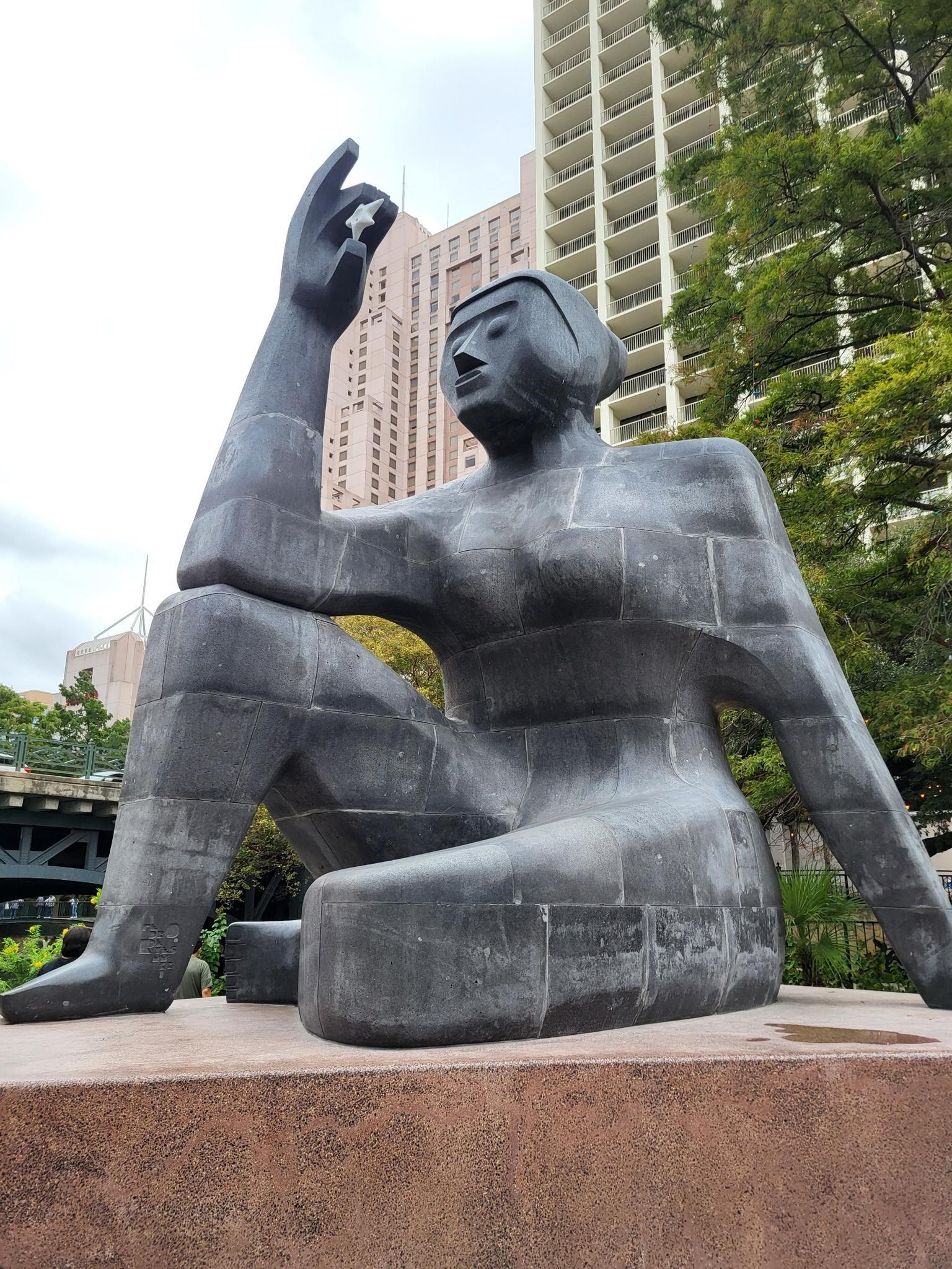 Large blocky statue of a seated person with a raised hand in an urban setting.