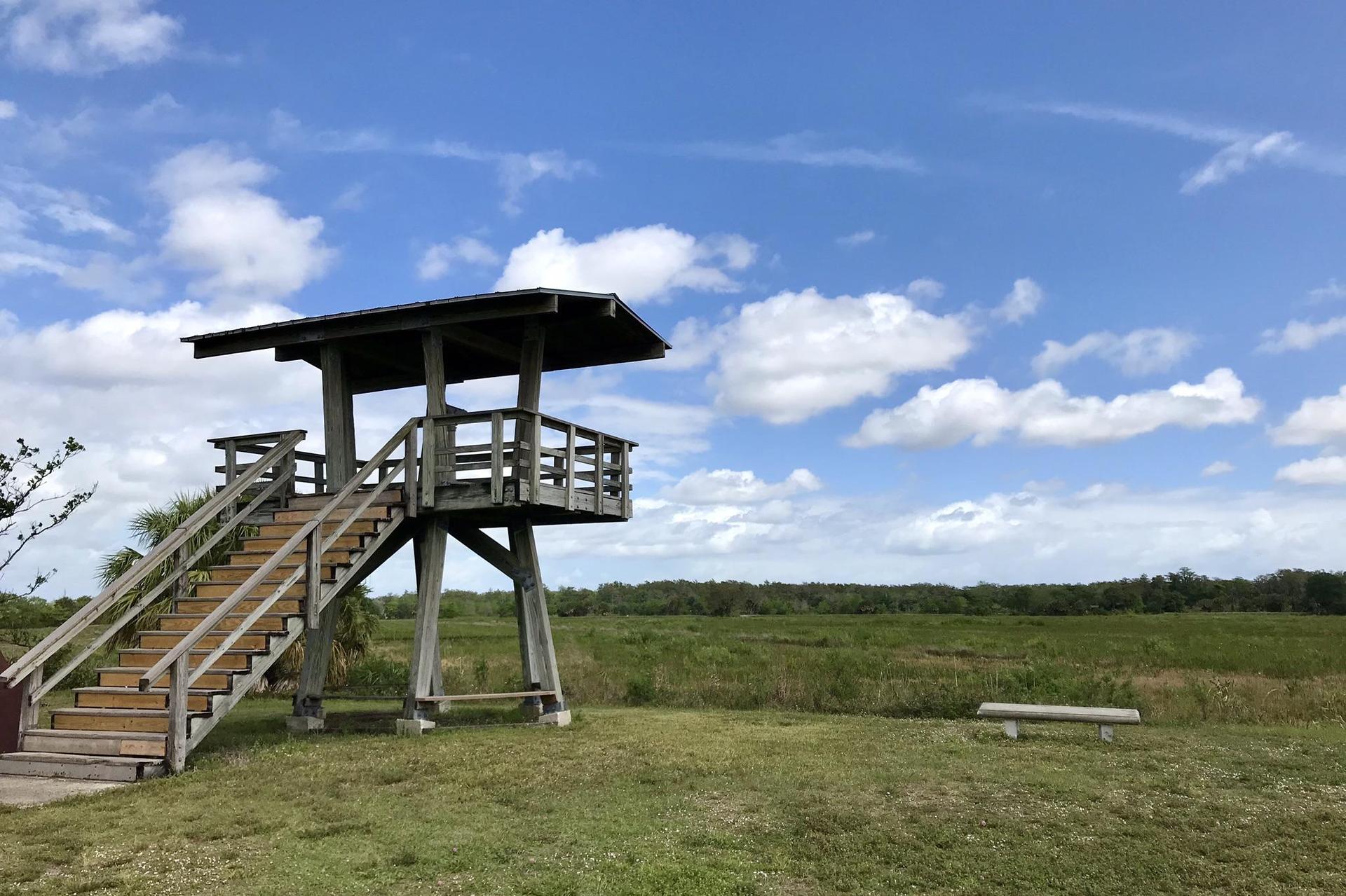 Wooden observation tower in a sunny grassy field under a cloudy sky.