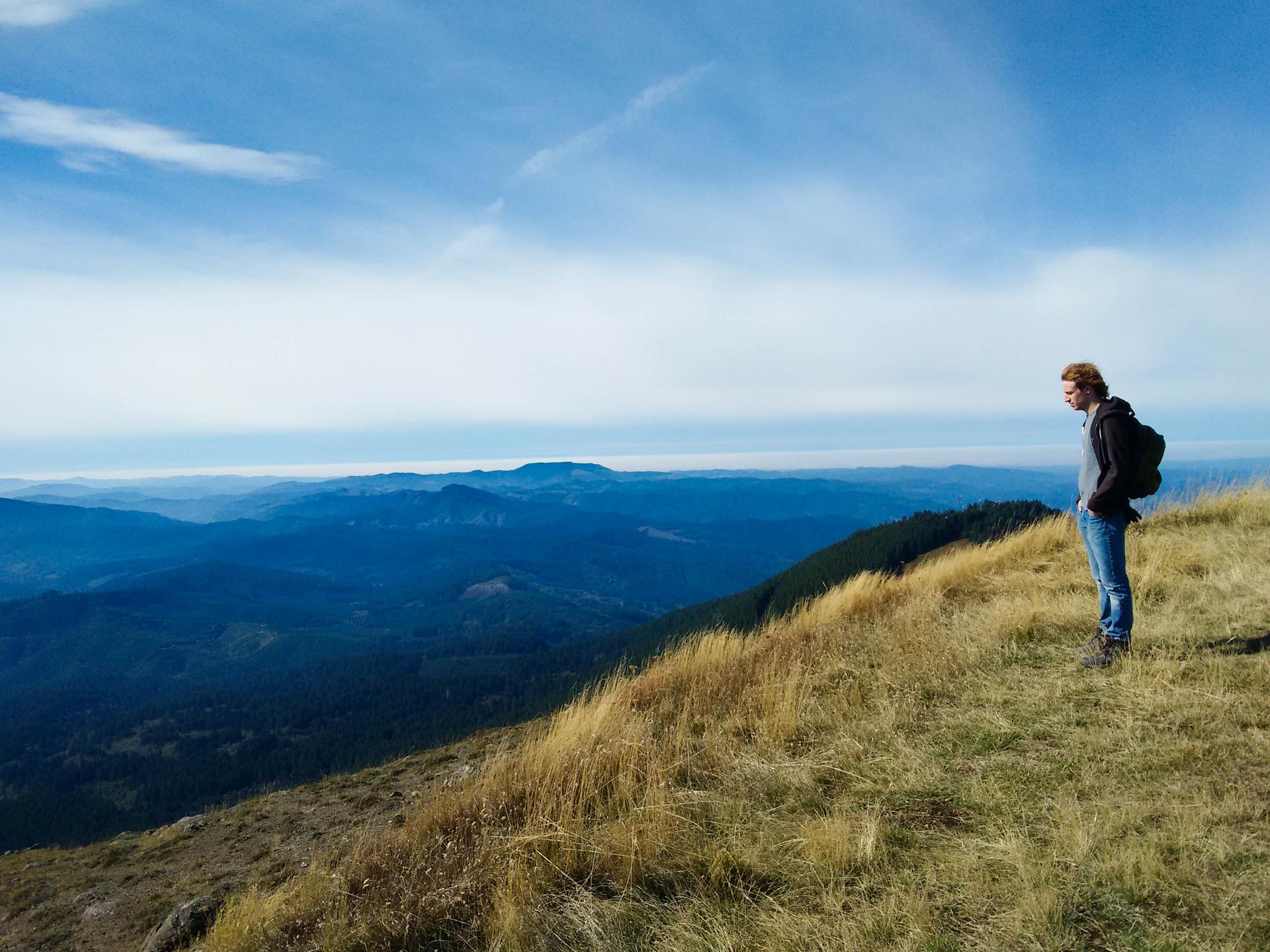 Man with backpack stands on a sunny grassy ridge overlooking blue mountains.