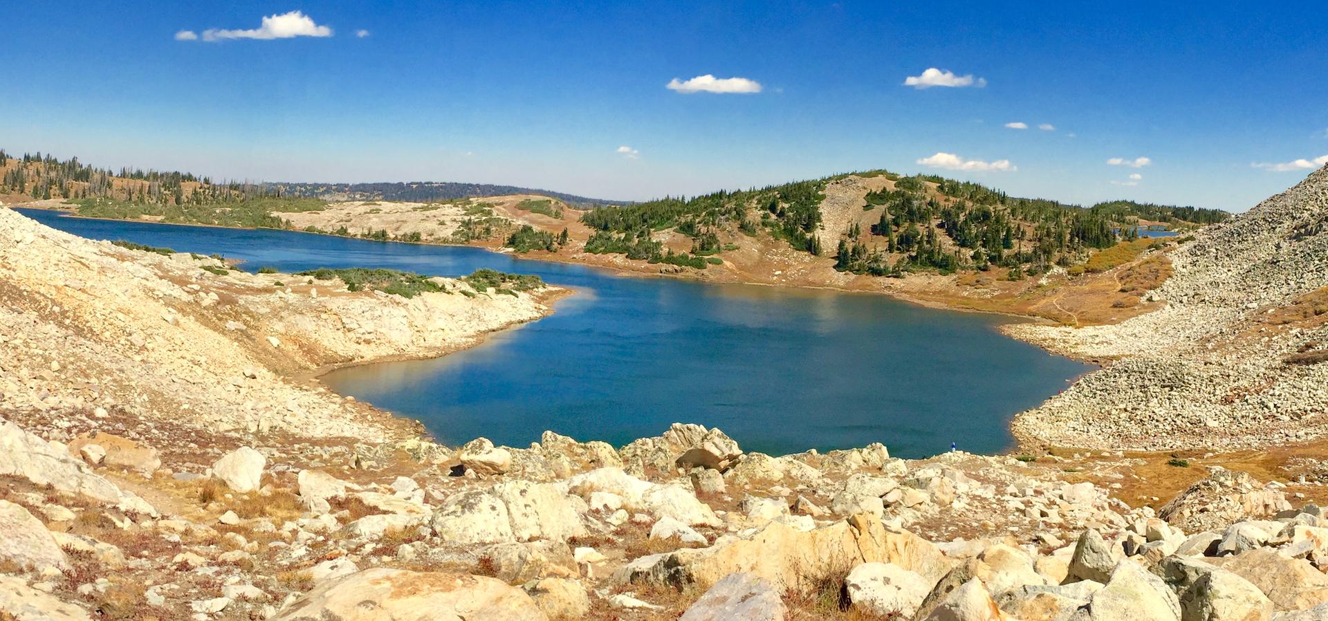 Blue lake surrounded by dry rocky hills and distant green trees.