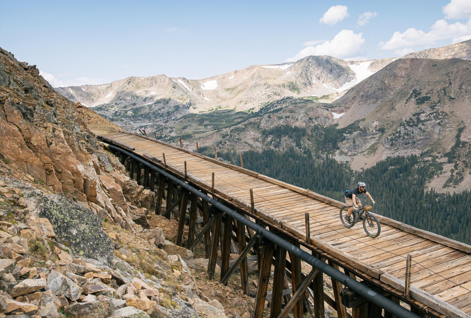 Mountain biker rides wooden trestle bridge over a vast mountain landscape.
