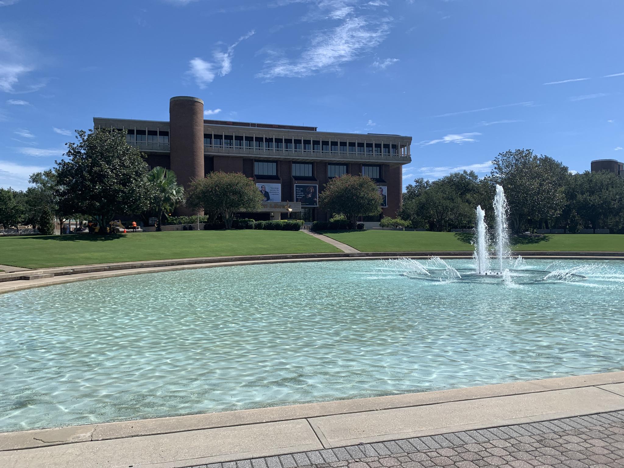 A large building behind a fountain in a pool, with green lawns and trees.