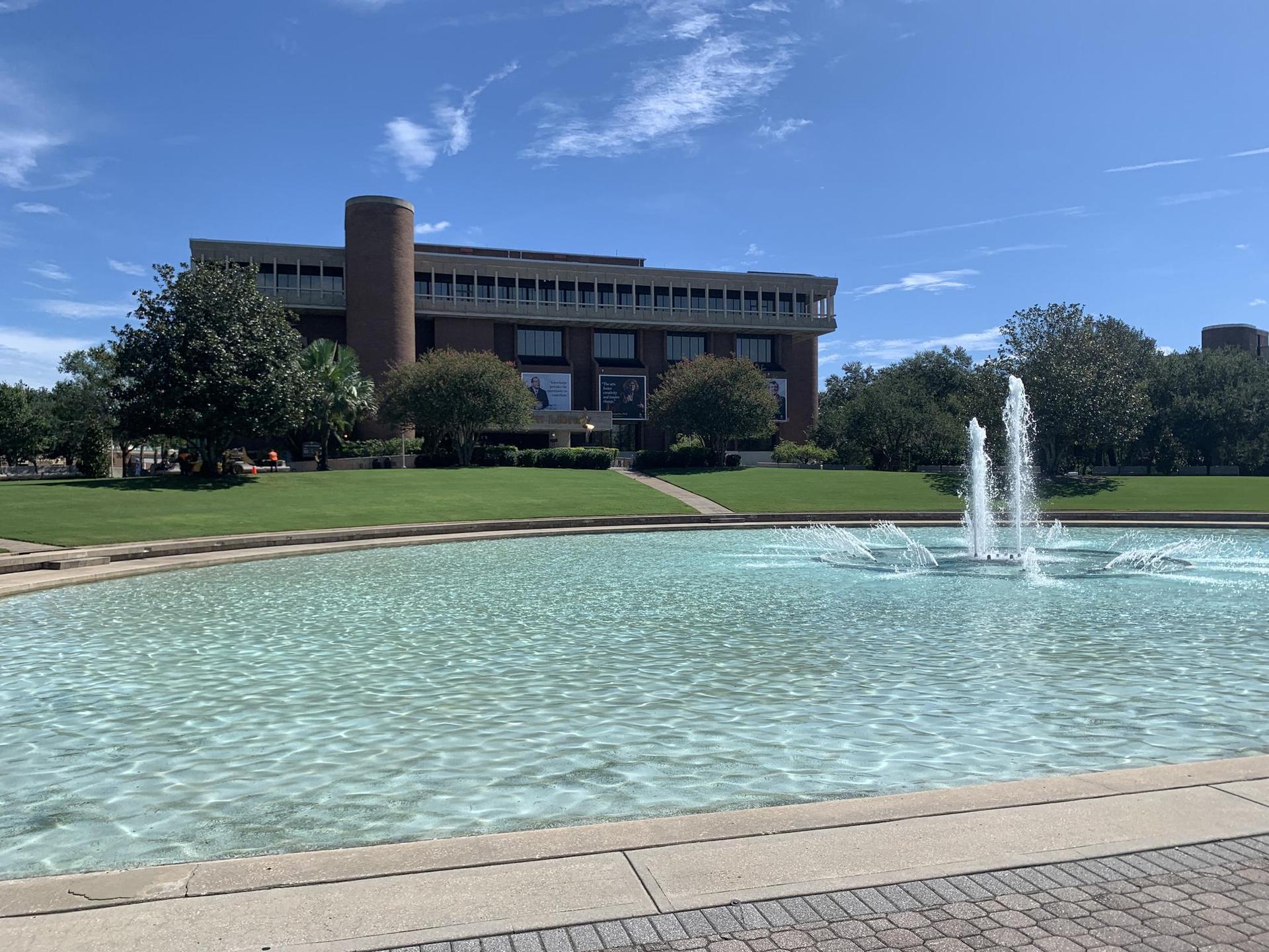 A large building behind a fountain in a pool, with green lawns and trees.