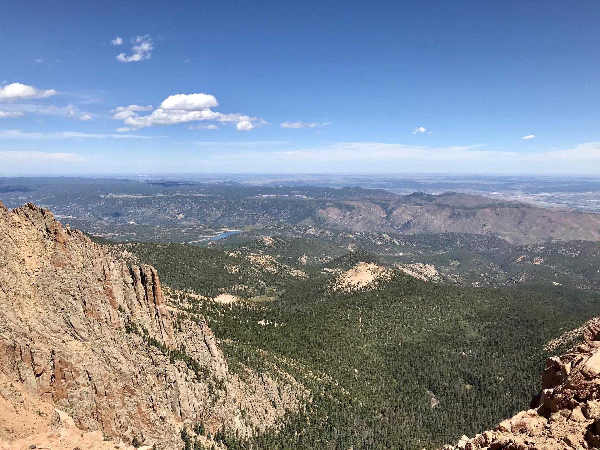 Expansive mountain landscape with forests and distant water under blue sky.