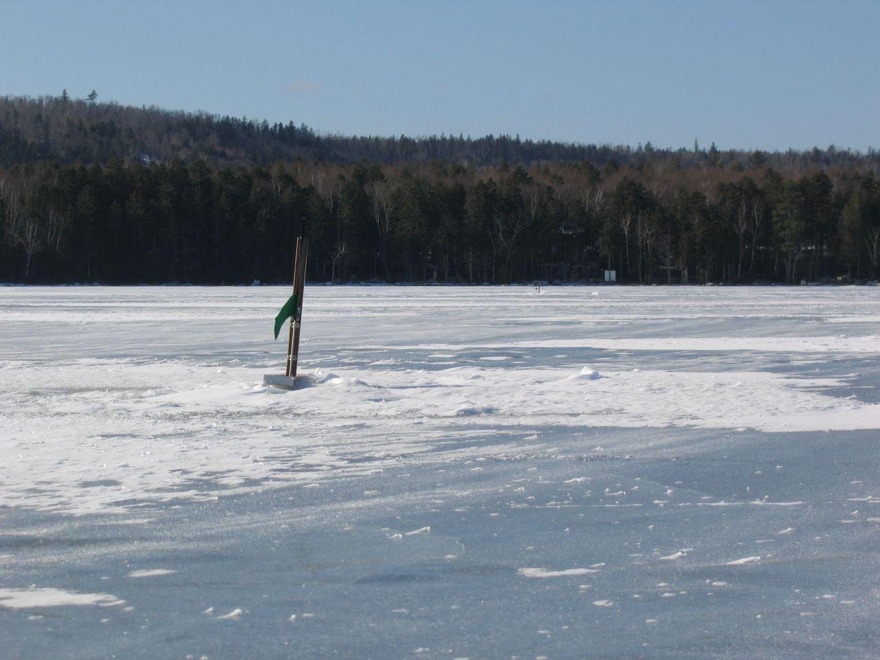 A green flag marks an ice fishing hole on a vast frozen lake.