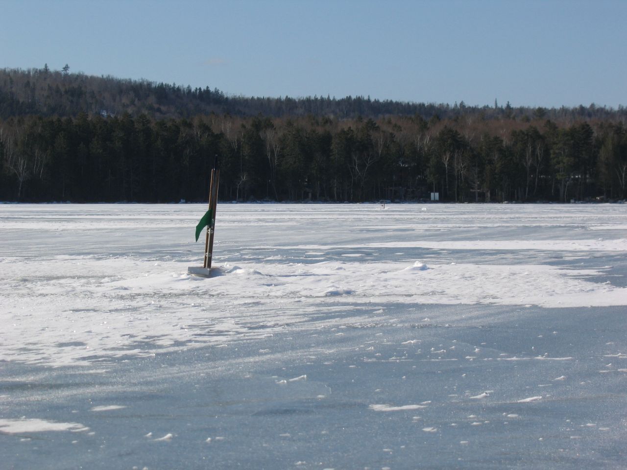 A green flag marks an ice fishing hole on a vast frozen lake. - Park Tote Road from Togue Pond to South Branch Pond — Gravel