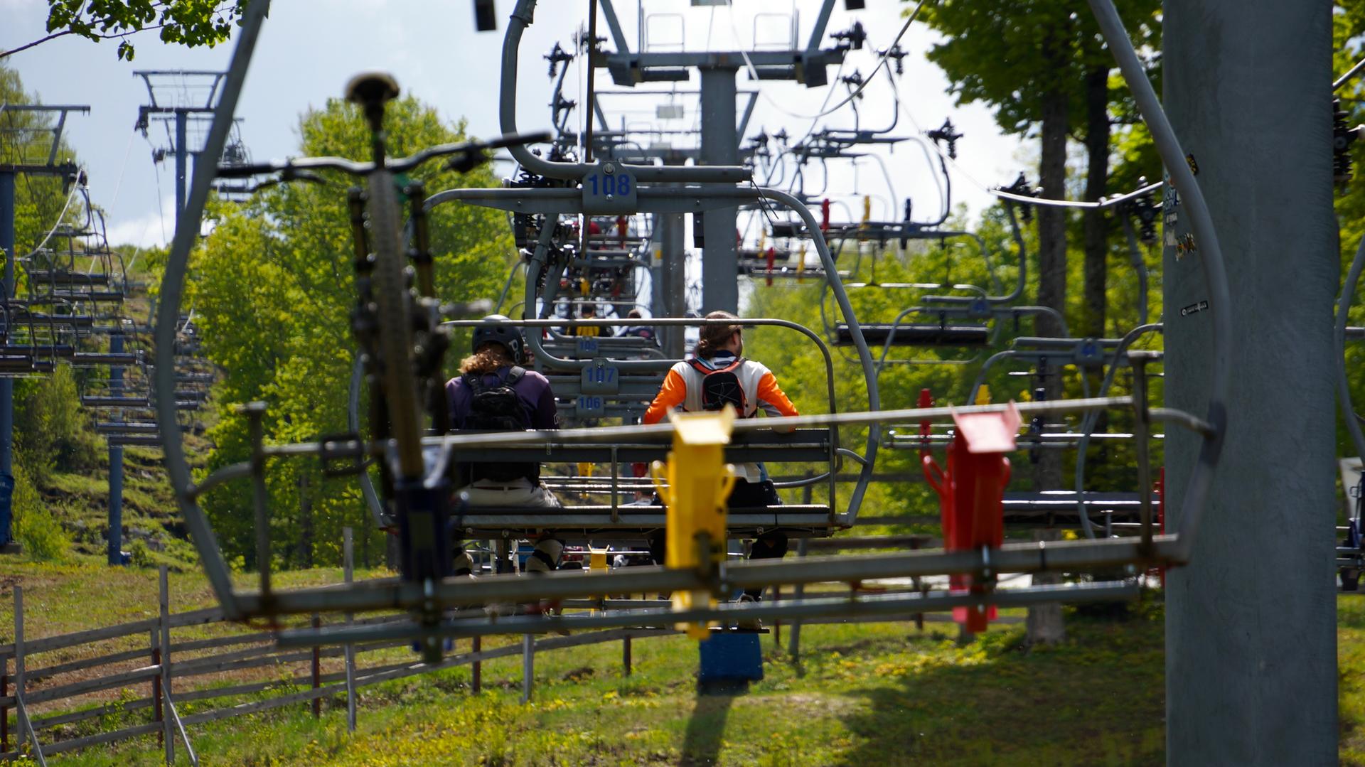 Two people ride a ski lift with mountain bikes on a sunny day.
