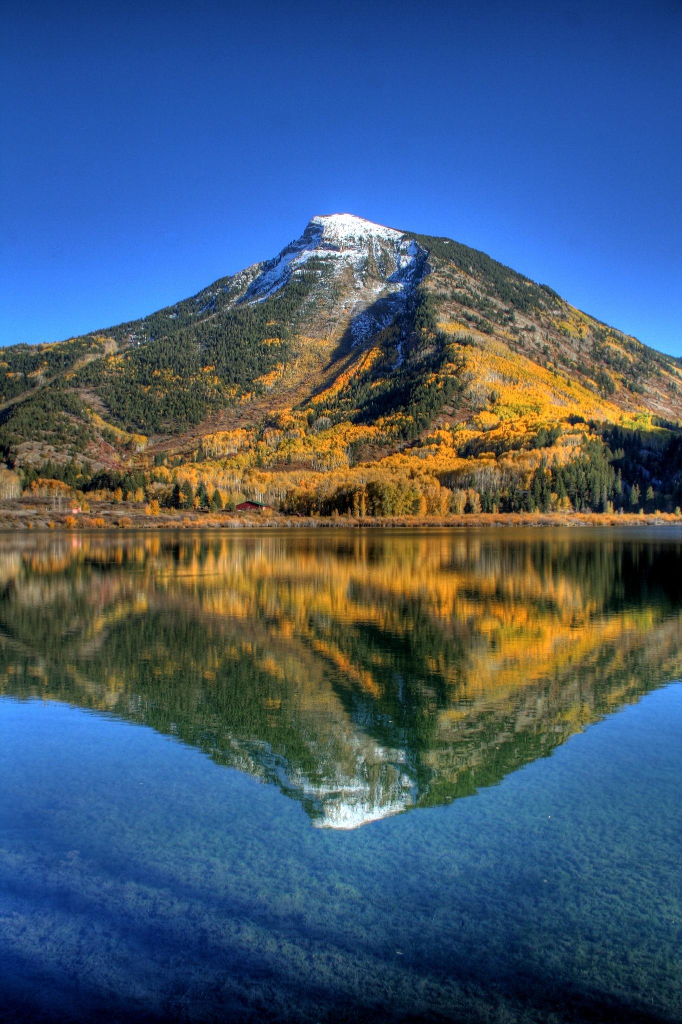 Snow-capped mountain with golden autumn trees reflecting in a clear blue lake.