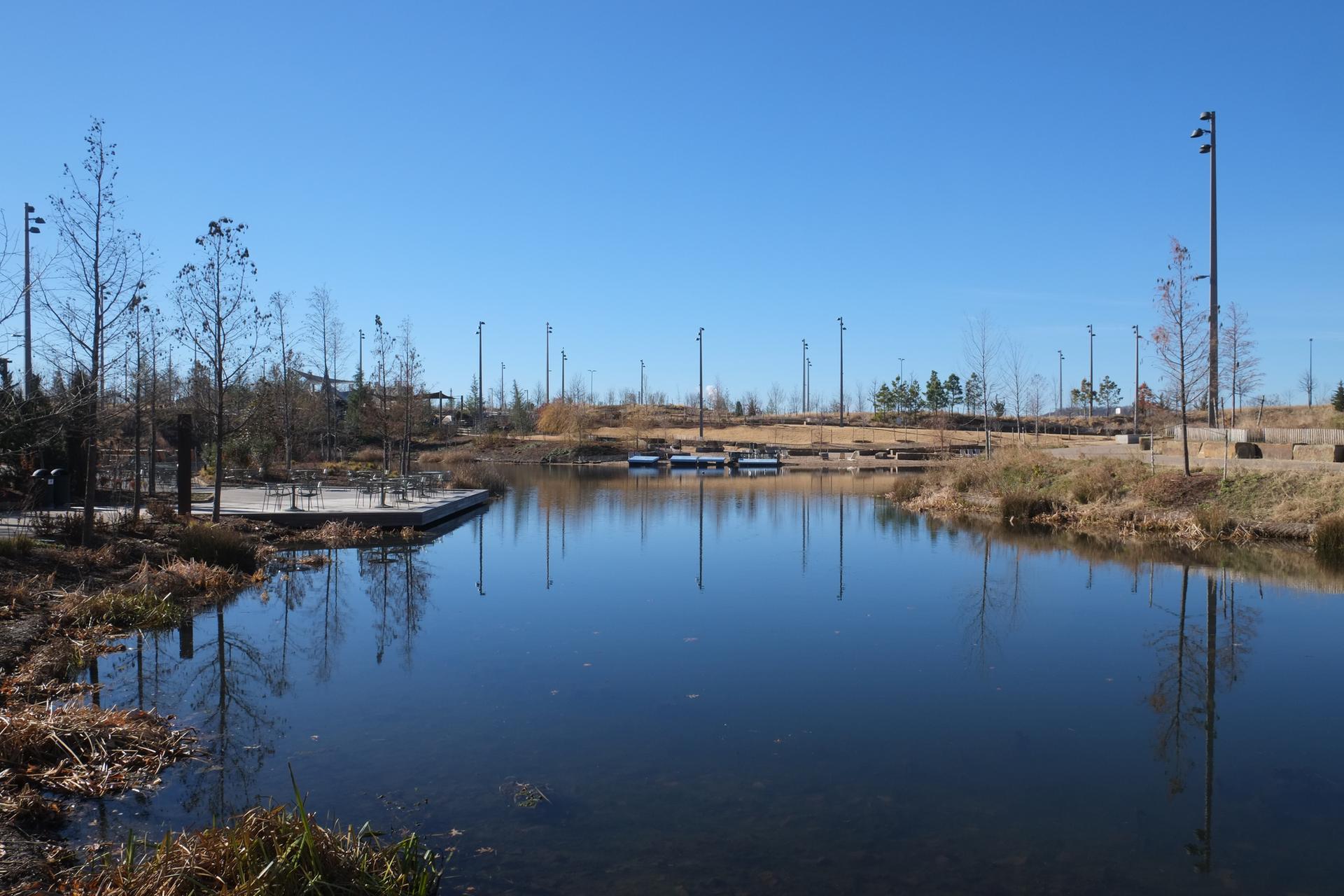 Calm pond with docks and tall lights, surrounded by bare trees under a blue sky.
