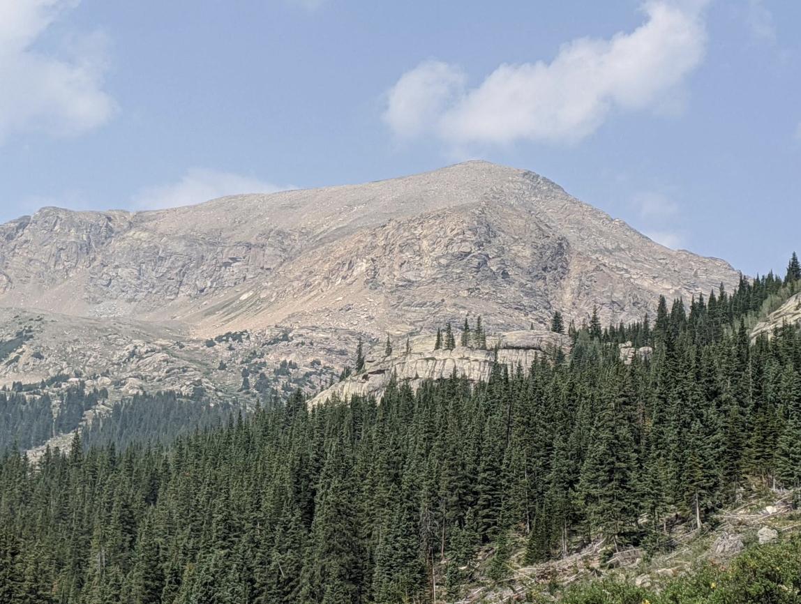 Rocky mountain peak with evergreen forest below, under a partly cloudy sky.