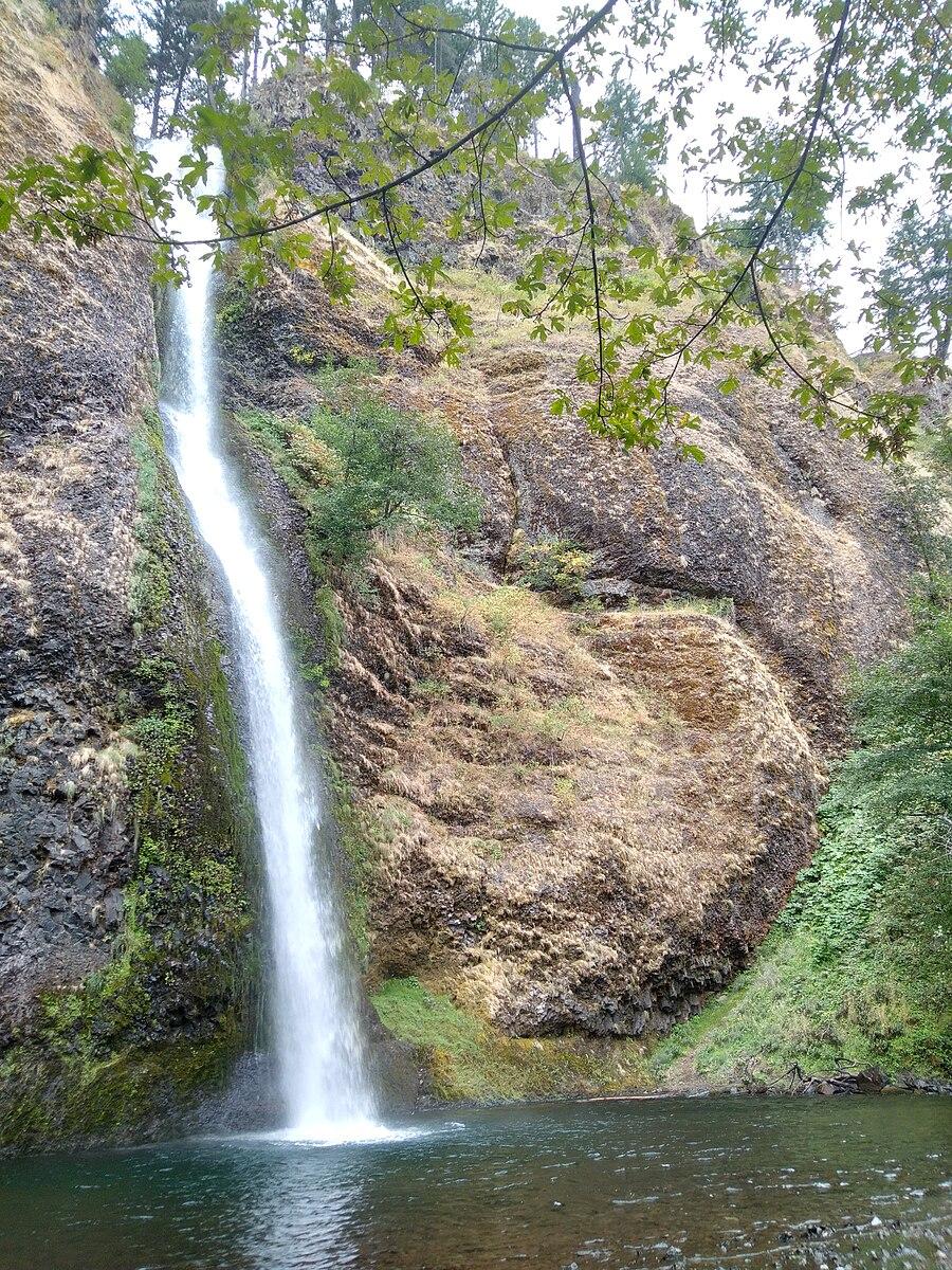 A tall, narrow waterfall cascades down a rocky cliff into a pool.