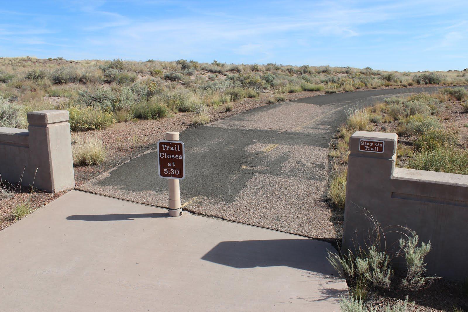 Paved trail entrance with "Trail Closed" sign in a dry, grassy landscape.