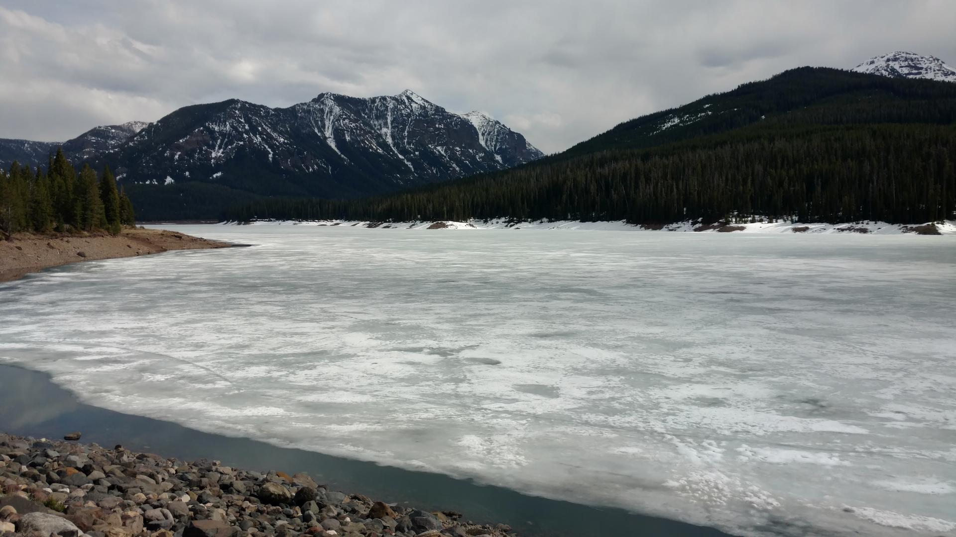Partially frozen lake with snow-capped mountains and evergreen forests under an overcast sky.