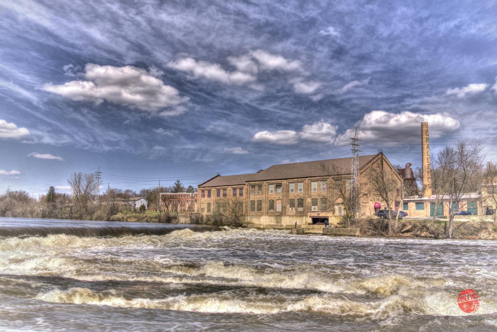 Old factory building by a turbulent river under a dramatic cloudy sky.