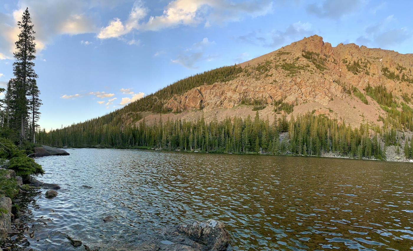 Serene mountain lake with a rocky shore, dense pines, and sunlit peak.