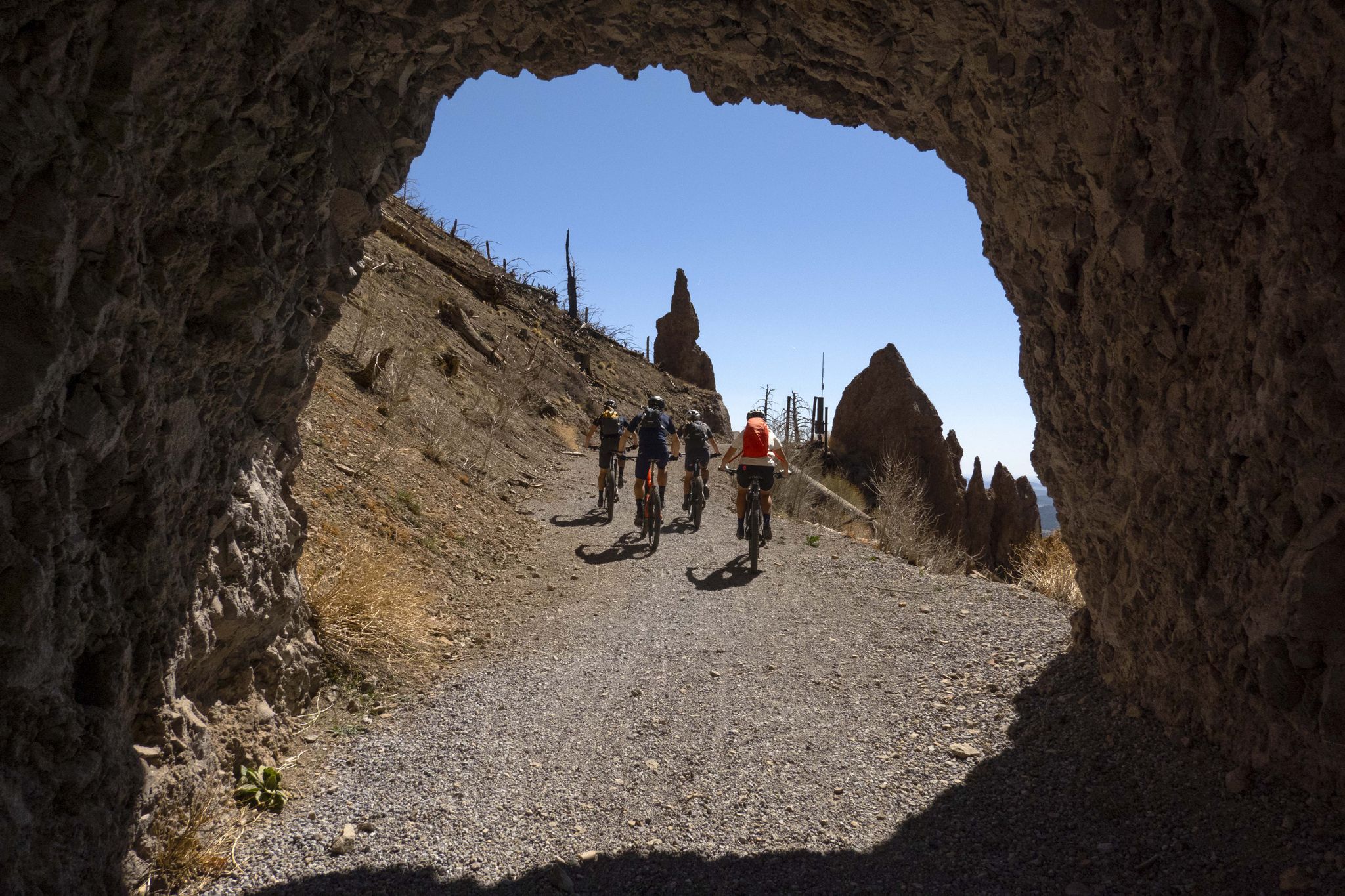 Cyclists emerge from a dark rock tunnel onto a sunny path with rock formations. - Lockett Meadow via Waterline Road