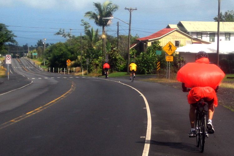 Three cyclists ride on a sunny tropical road with houses and palm trees. - Volcano to Keaʻau loop