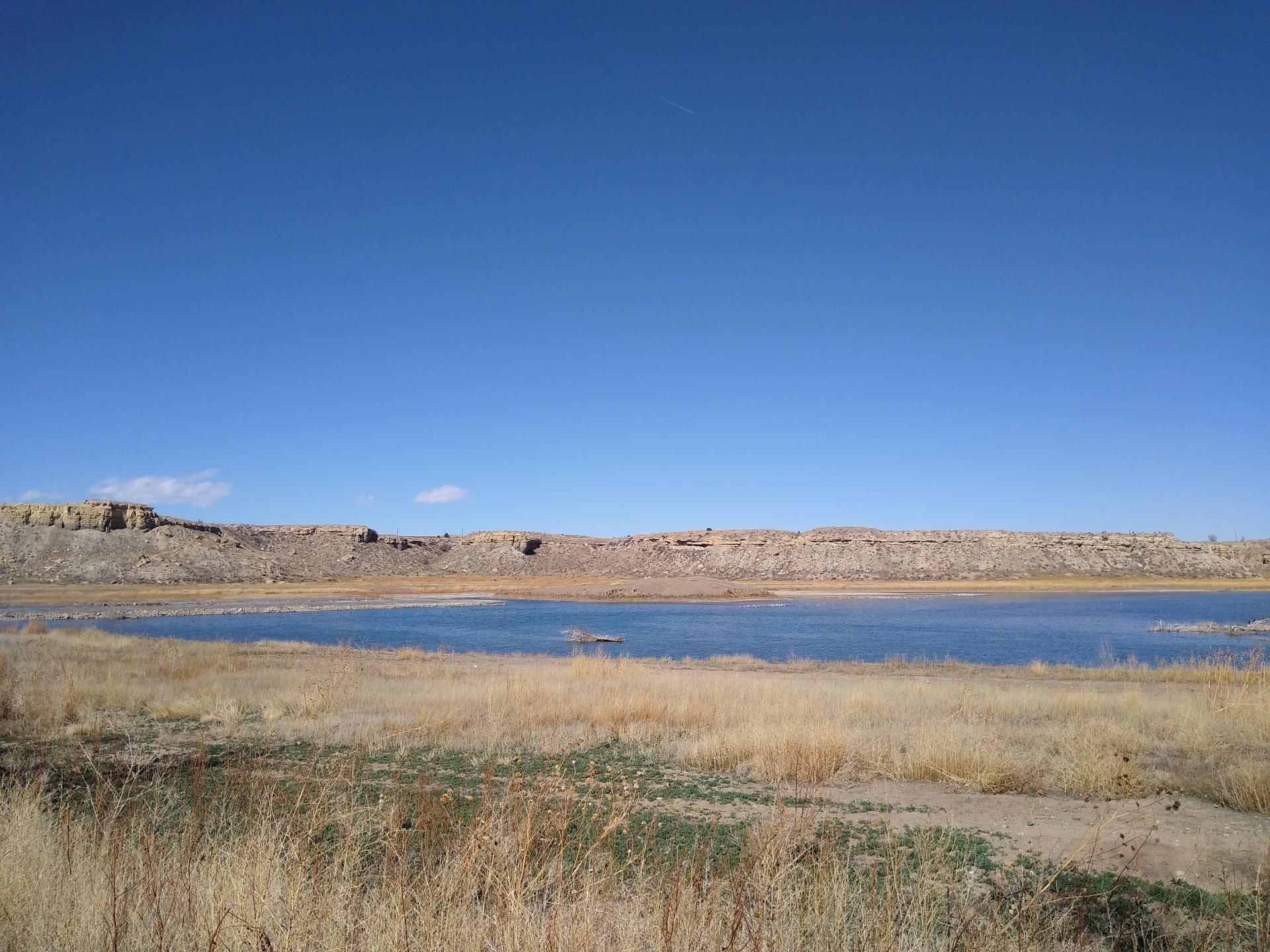 A wide lake with dry grass in the foreground and rocky hills under blue sky.