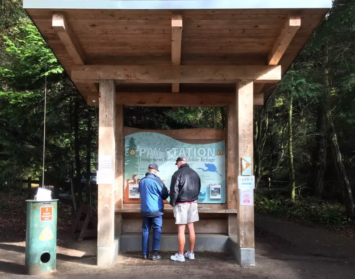 Two men stand at a wooden pay station in a forested area.