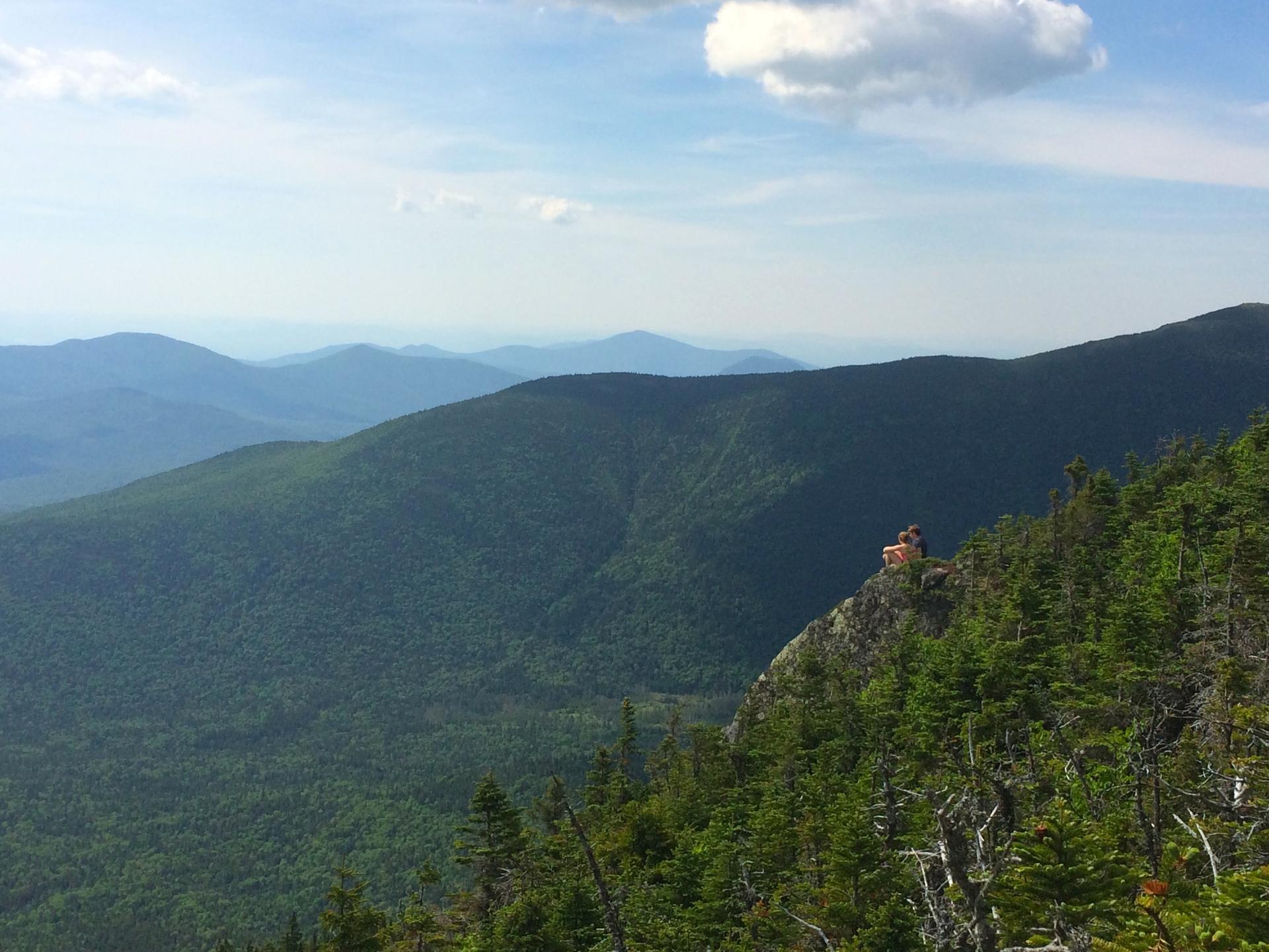 Person sits on a rocky cliff overlooking vast green mountain ranges.