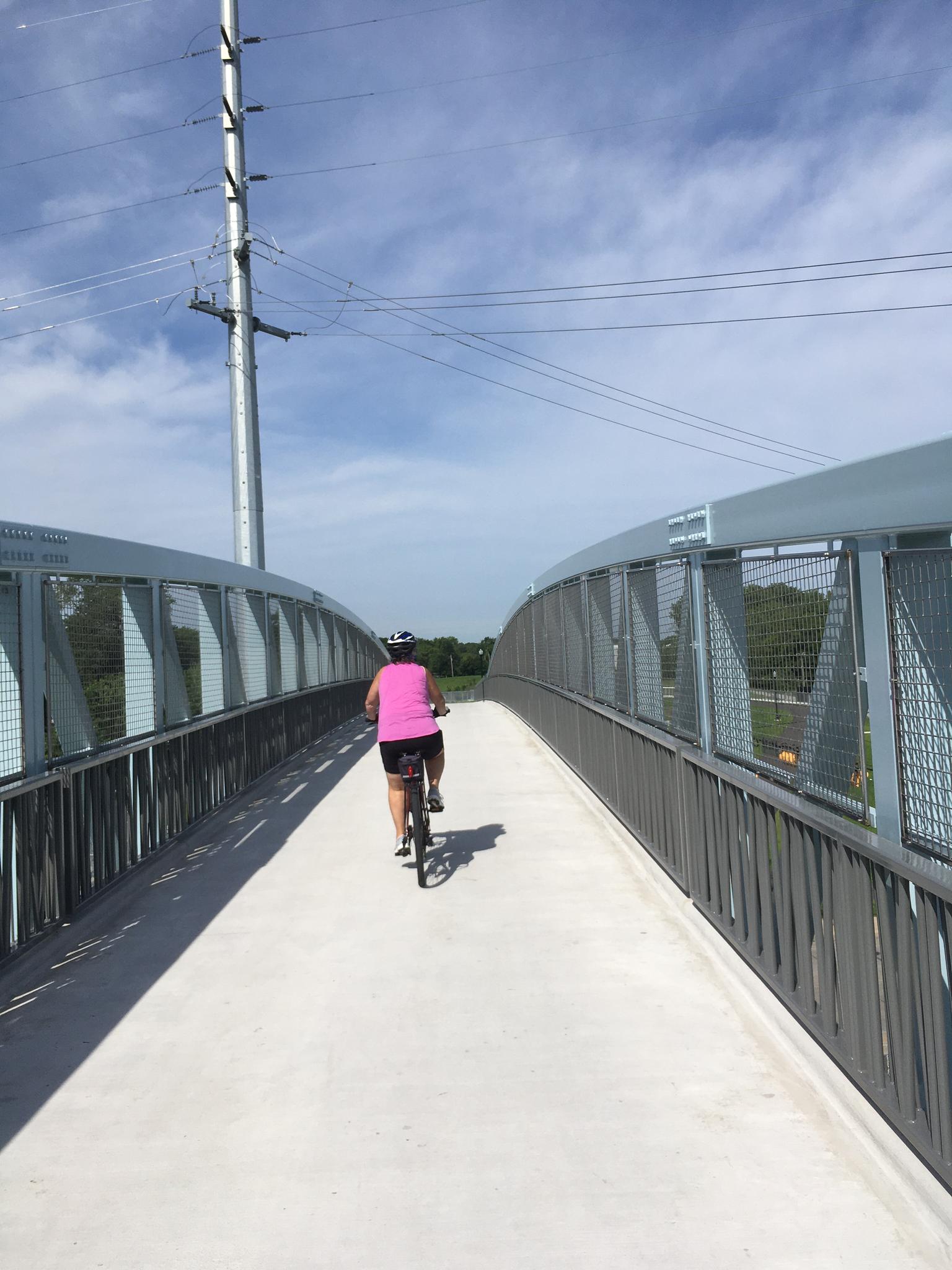 Cyclist in pink shirt on a modern metal bridge under a bright sky.