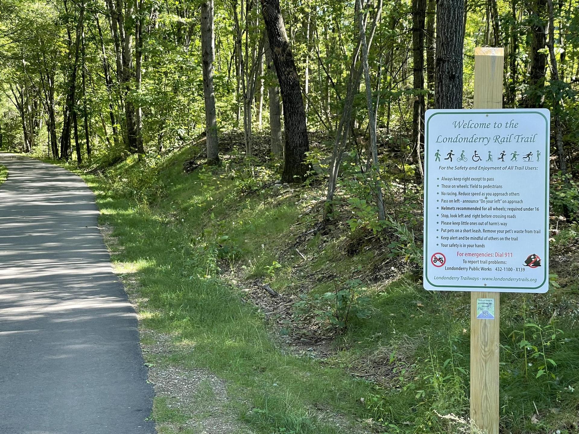 Paved path through a sunny forest with a trail information sign.
