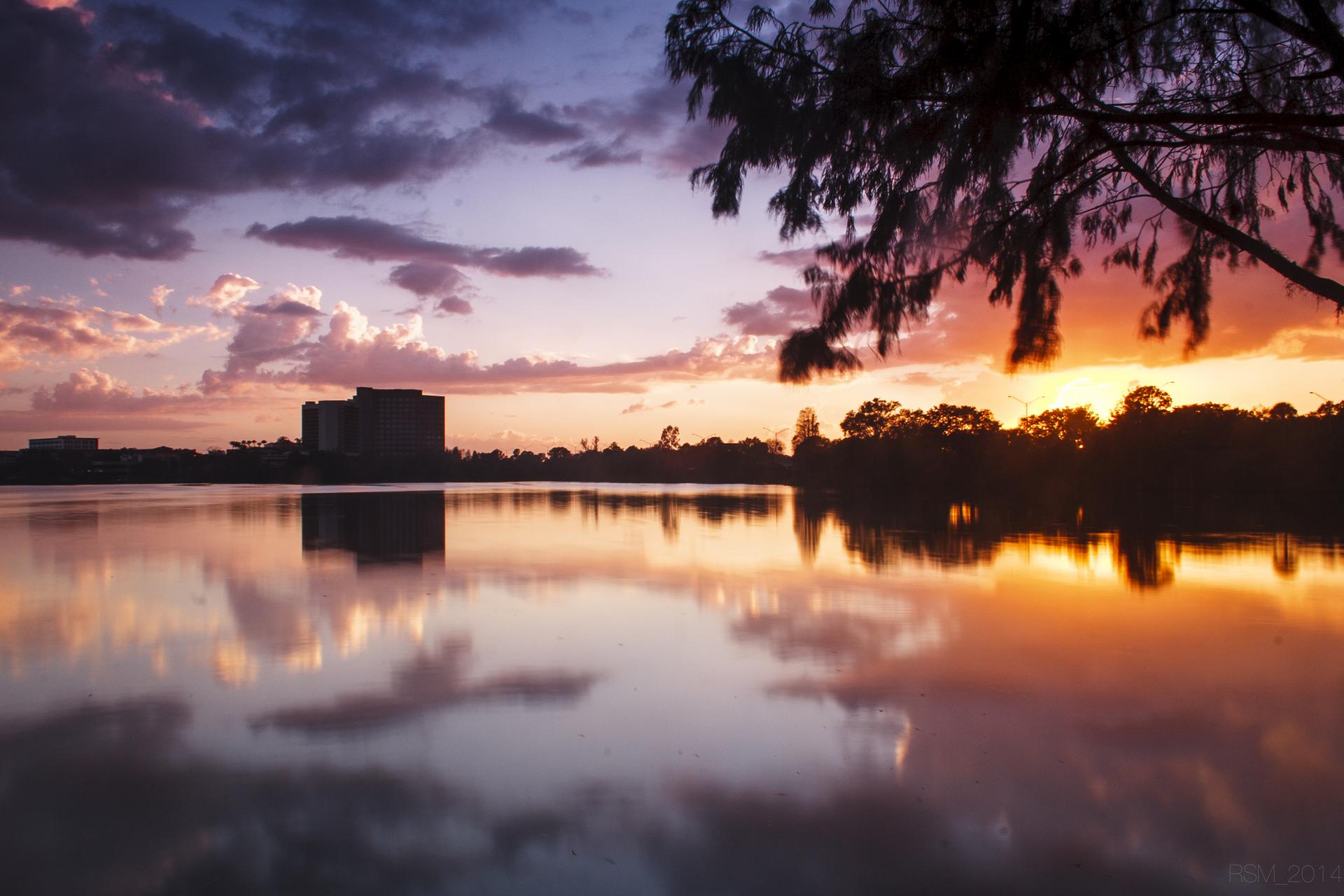 Vibrant sunset over a calm lake, reflecting colorful clouds and distant buildings.