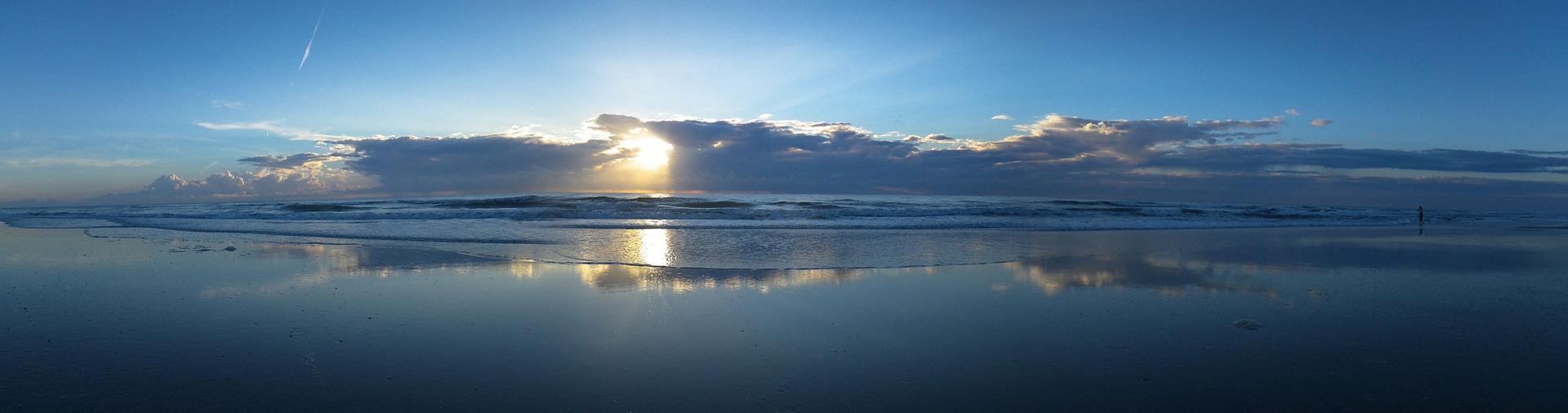 Sun peeking through clouds over a calm ocean, reflecting on wet sand.