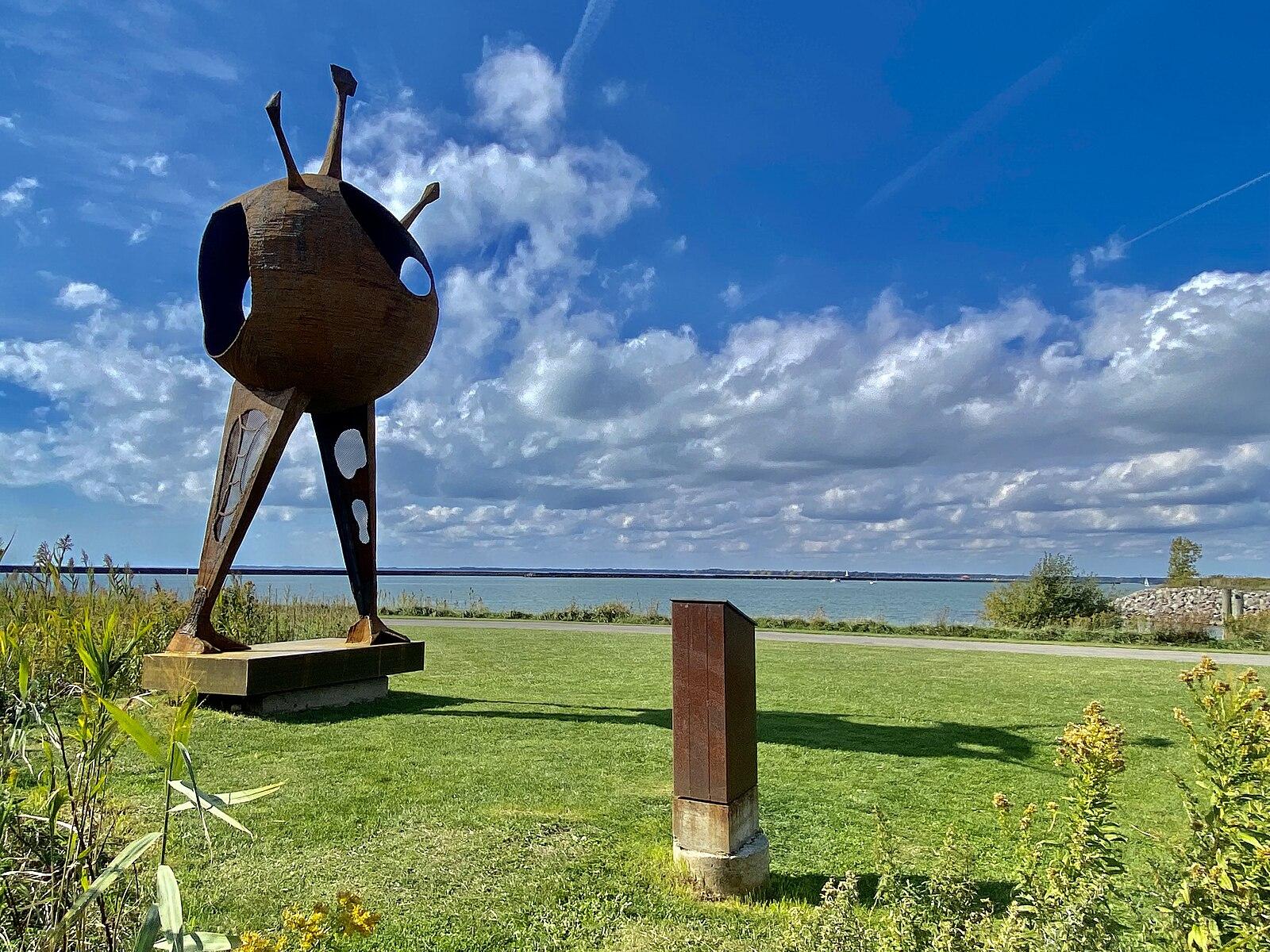Large abstract sculpture on green grass by water under blue sky.