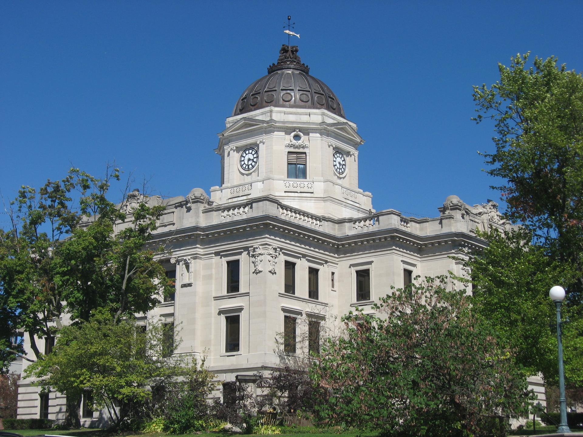 A grand stone building with a large dome and clock tower, surrounded by green trees under a clear blue sky.