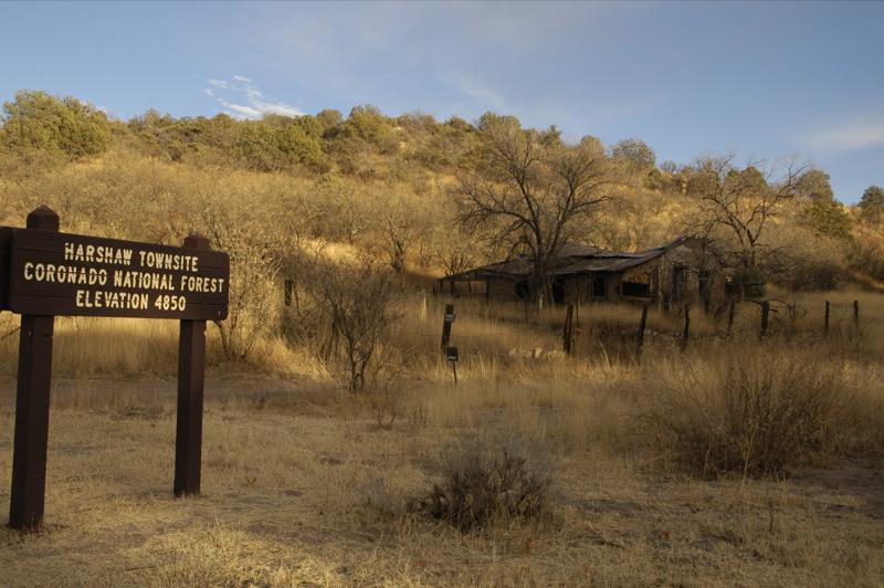 A wooden sign stands in a dry field near an old, dilapidated building.