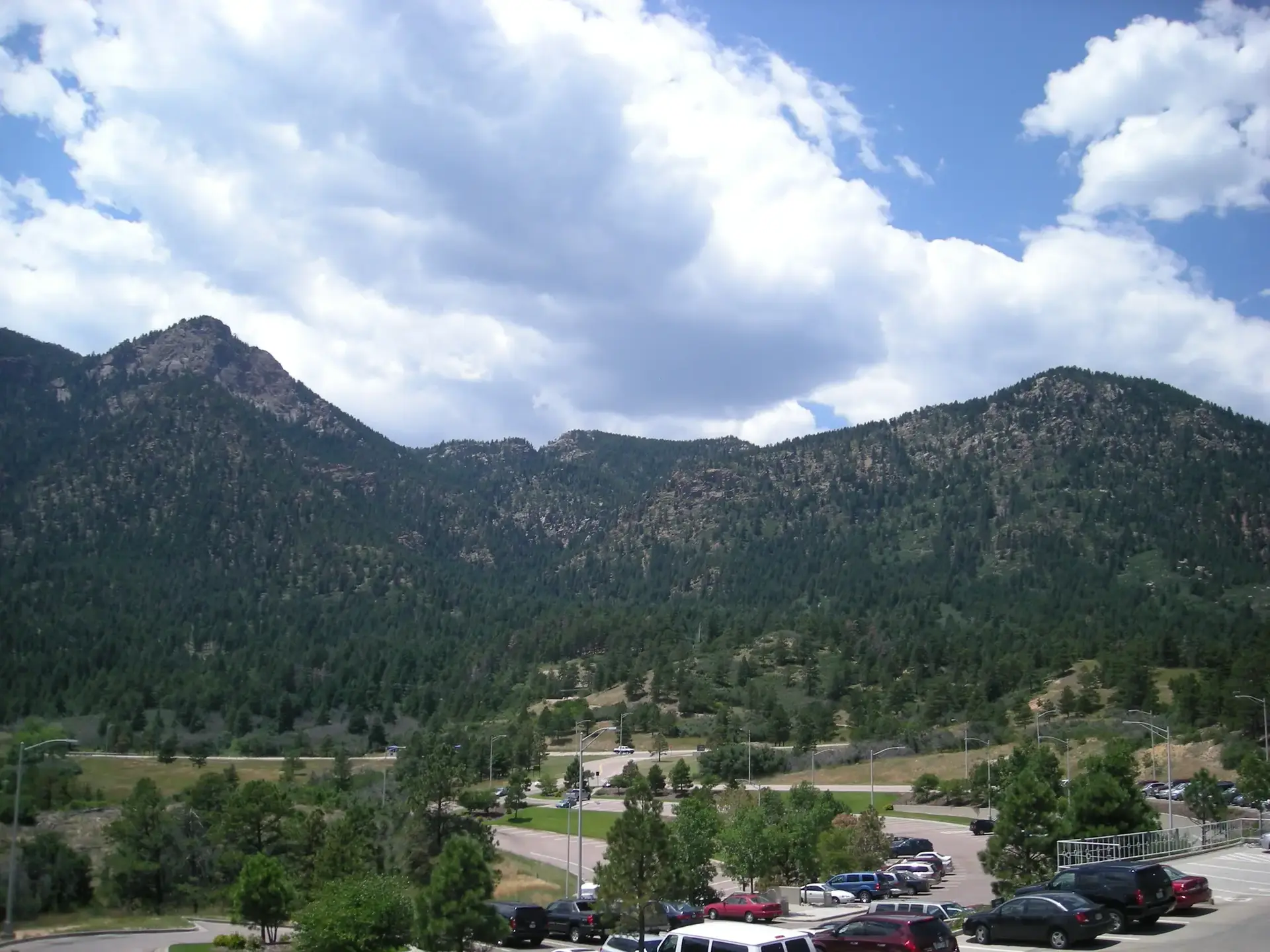 Tree-covered mountains overlook a valley with roads and busy parking lots under a cloudy sky.