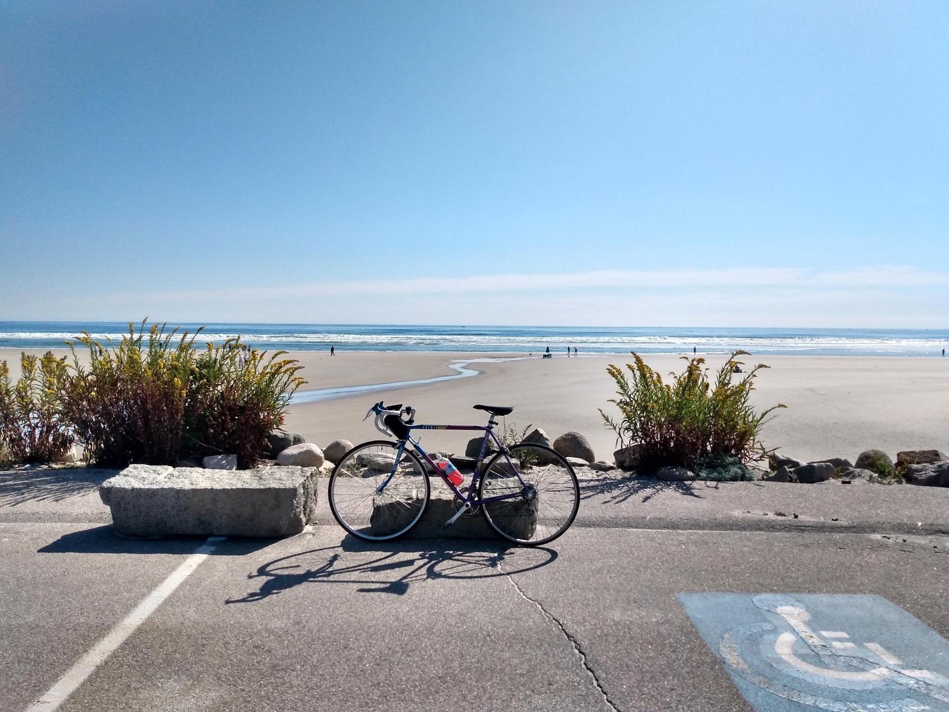 Bicycle parked on sunny beach pavement with ocean waves and distant people.