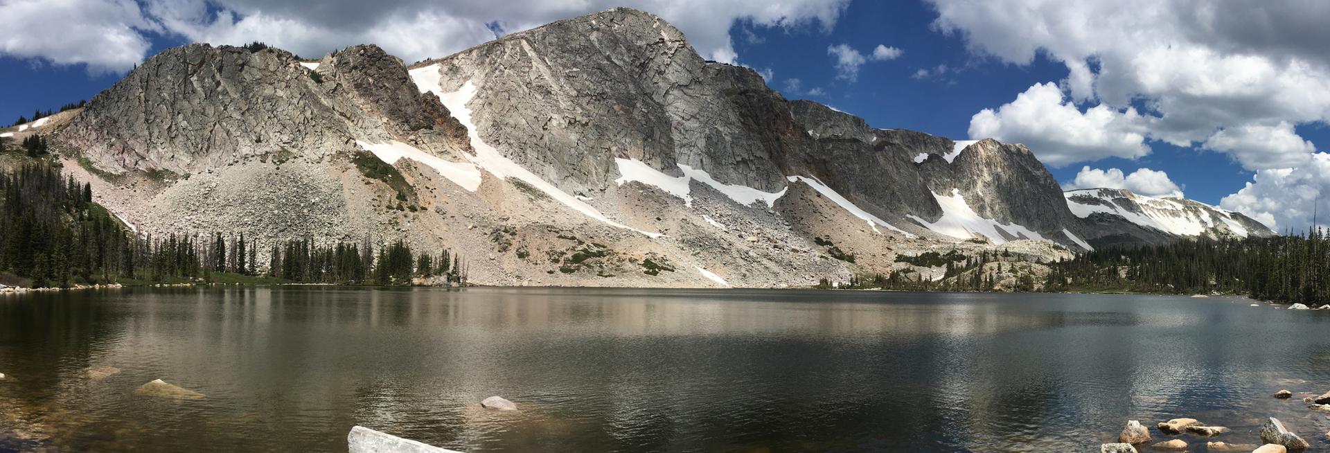 Panoramic view of a serene mountain lake reflecting snow-capped peaks under a blue sky.