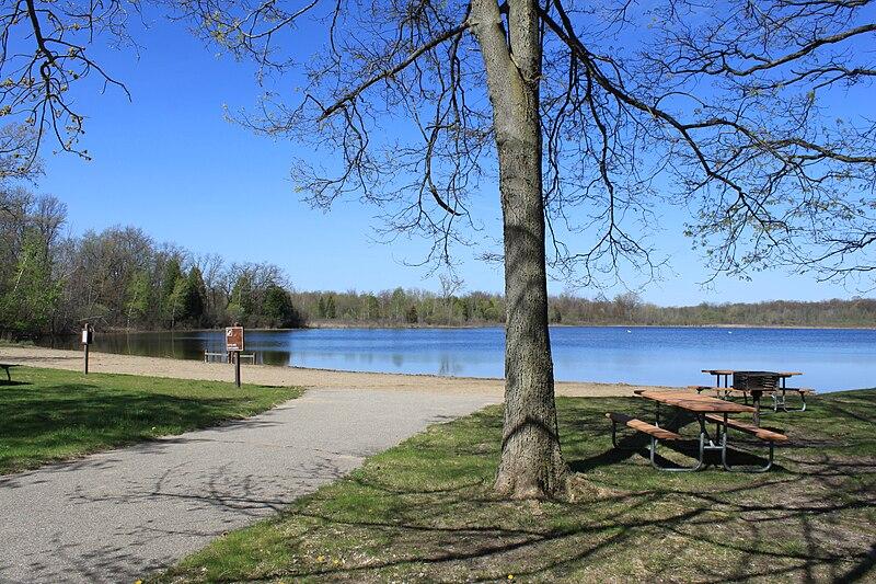 Picnic tables and a path by a calm lake on a bright, sunny day.