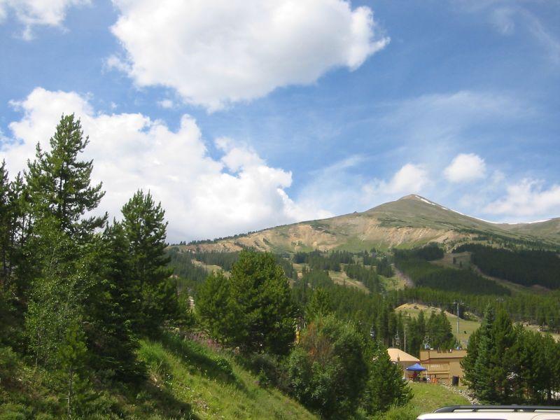 Green mountain slopes with pine trees and a building under a bright, cloudy sky.