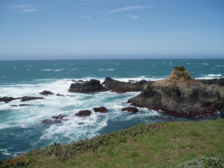 Sunny coastal view with crashing waves, rocky sea stacks, and green cliff.