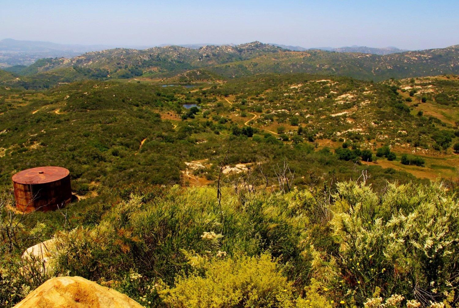 Rusty water tank in a vast, sunny, green, hilly landscape.