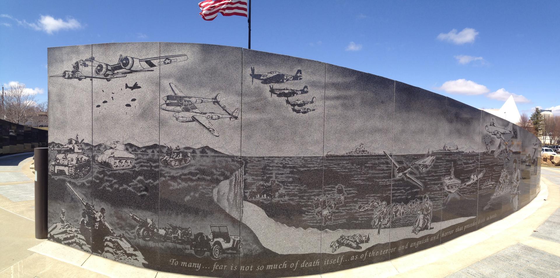 Curved stone wall with detailed engravings of historical war scenes, flag above.