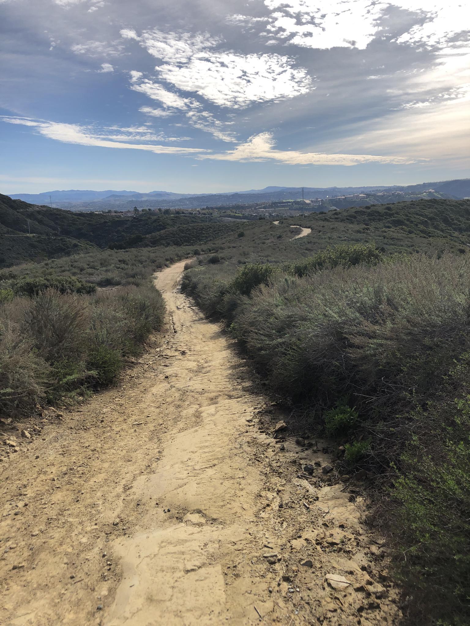 A winding dirt path through dry, brushy hills under a partly cloudy sky.