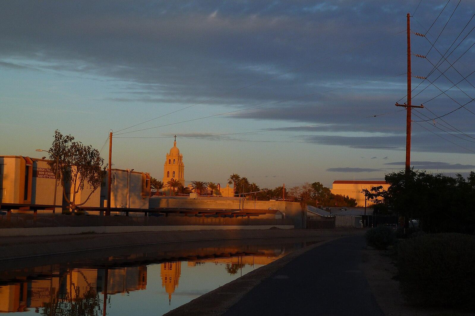 A canal reflects a distant building and power lines at sunset with cloudy sky.
