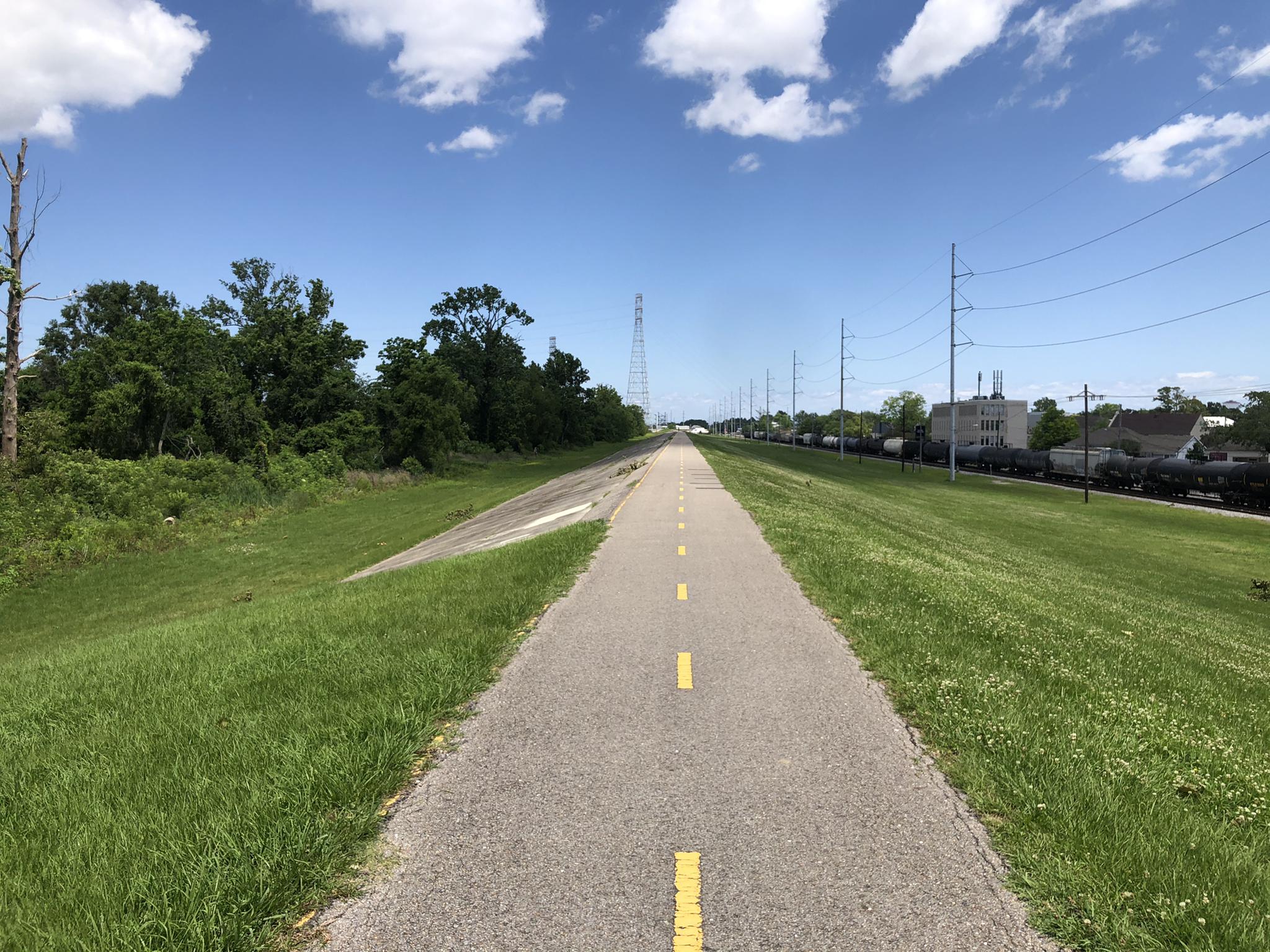 Long paved path through green grass, with power lines and distant train cars.