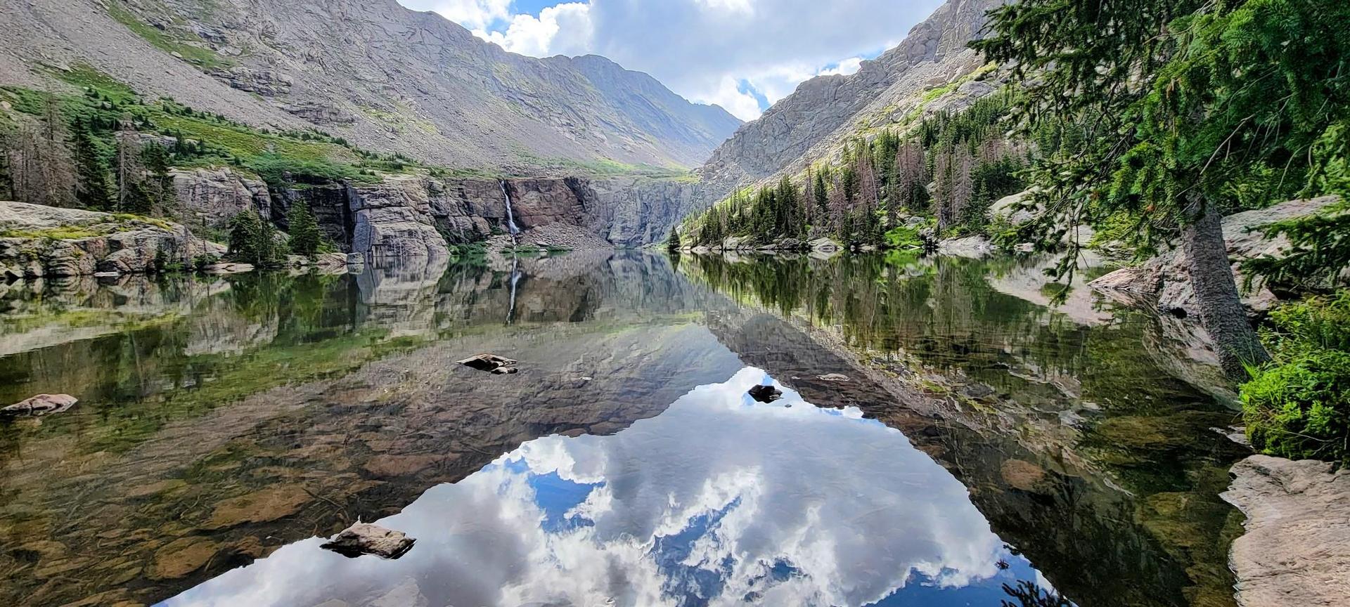 Calm mountain lake reflecting sky and peaks, with waterfalls in background.
