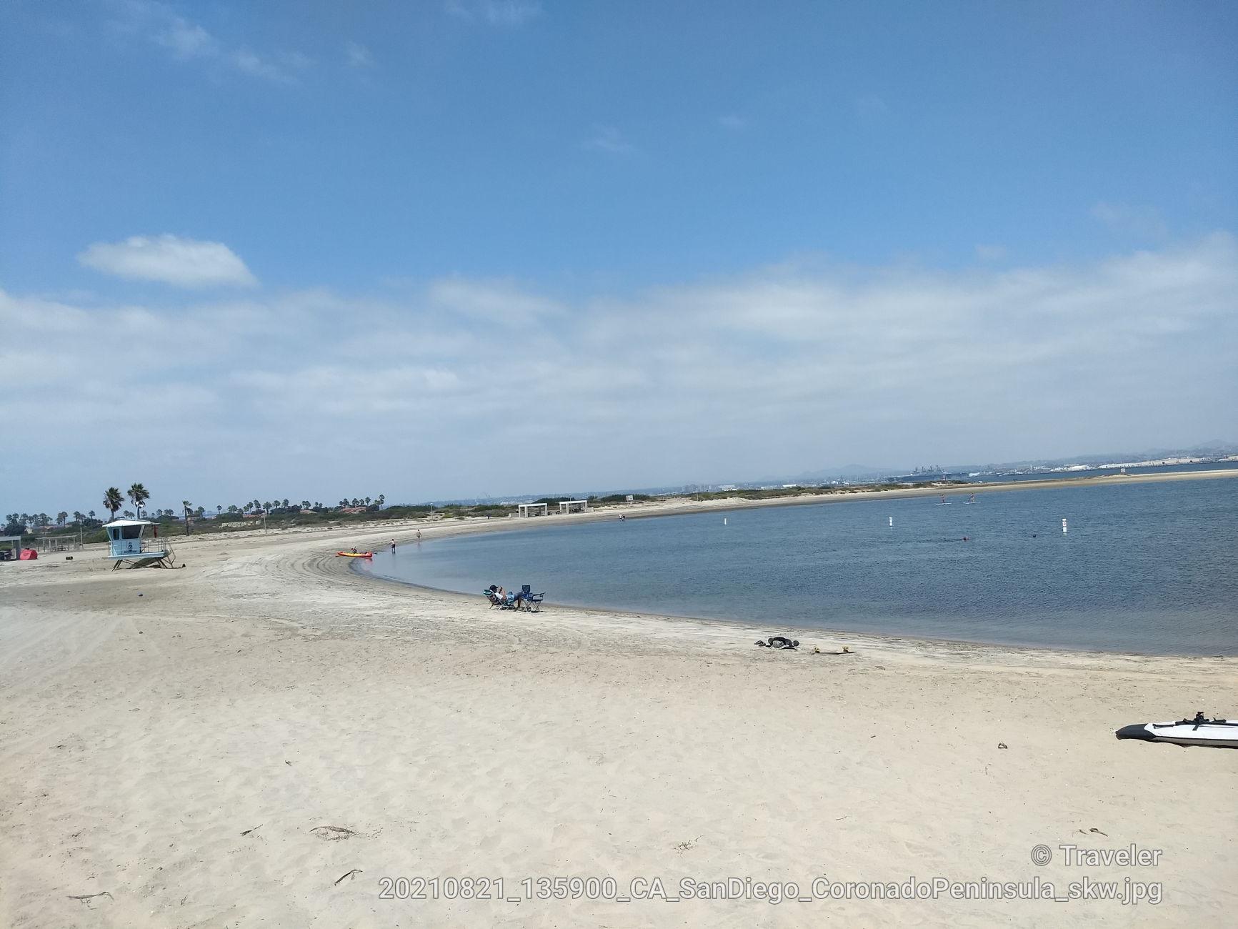 Wide sandy beach curves along calm bay water under a blue sky.